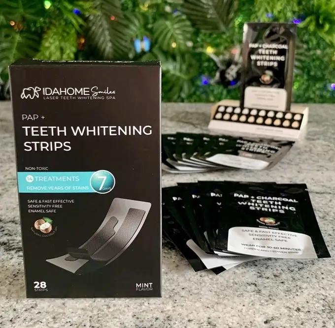 Close-up view of Idahome Smiles' Arctic Frost PAP+ Teeth Whitening Strips packaging, highlighting the product's non-toxic, enamel-safe qualities and the mint flavor. Visible in the background are the individually packaged strips, ready for use, and a tasteful display of additional Idahome Smiles products, all set against a greenery backdrop, emphasizing the brand's commitment to natural wellness and dental health.